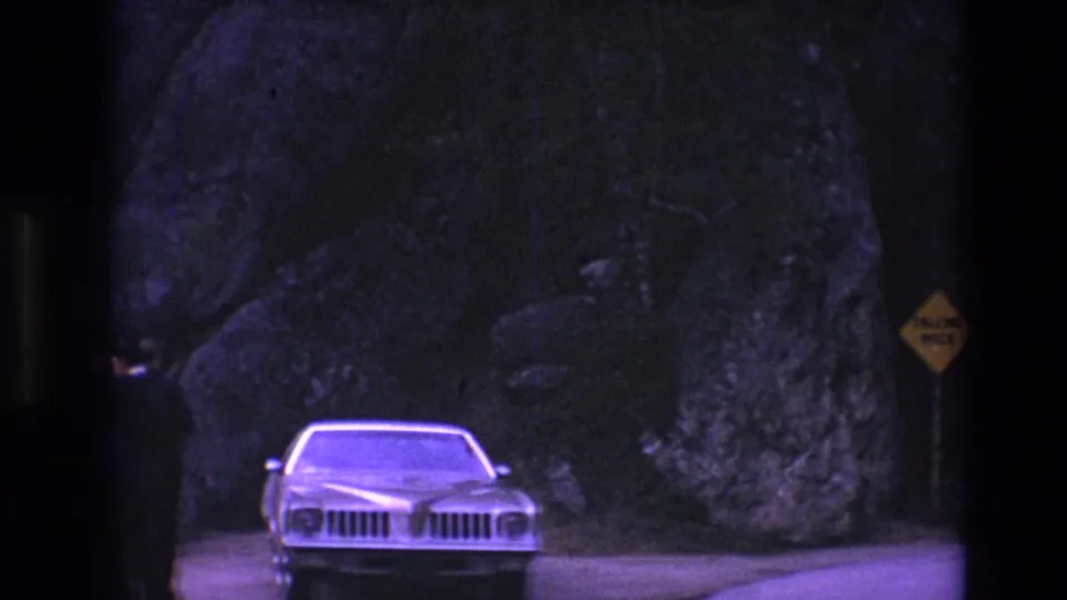 A light-colored car is stopped near a yellow falling-rock sign along a rocky South Dakota road in 1974 home-movie footage.