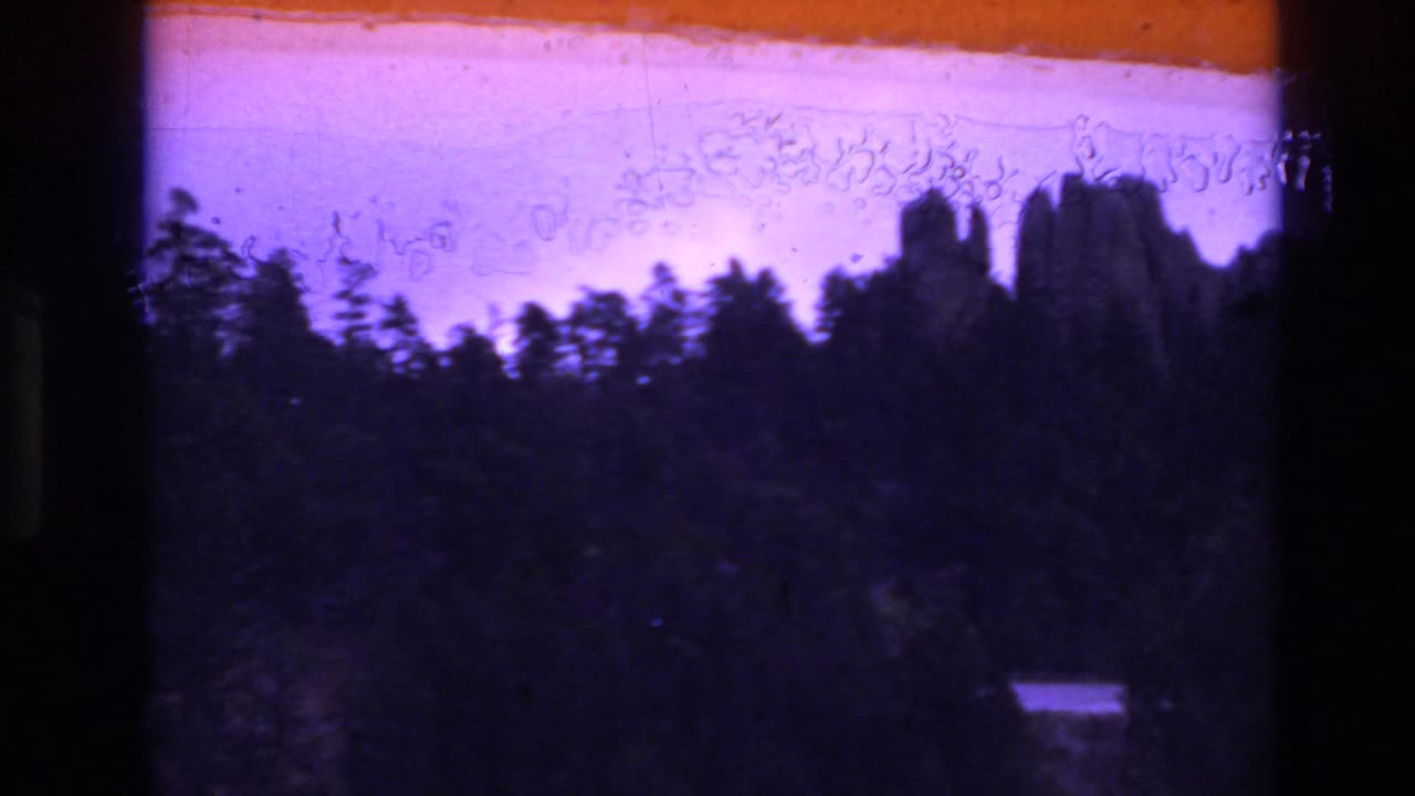 Faded 1974 South Dakota 8mm frame showing silhouetted rock formations behind trees, with a reddish-orange light leak and dark film borders.