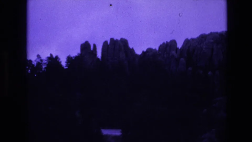 South Dakota travel footage frame with distant rock spires and a dark forested ridge silhouetted against a purple-blue faded sky.