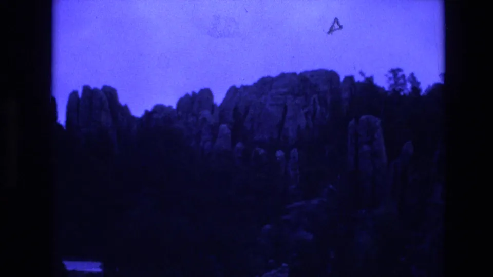 Blue-shifted 8mm frame from South Dakota showing rugged rock formations rising above a dark wooded foreground.