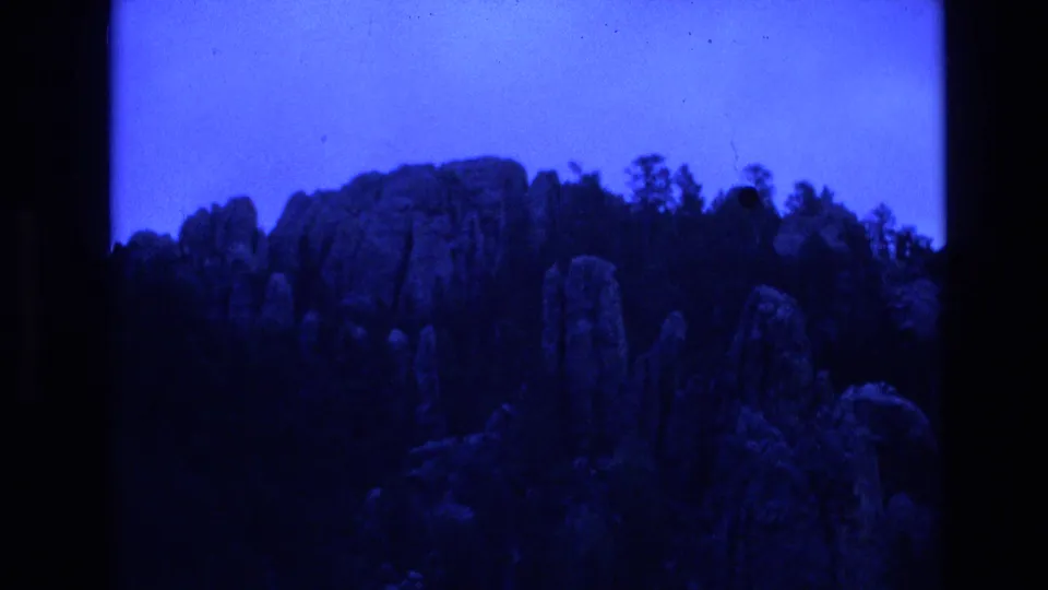 Faded 1974 South Dakota 8mm frame showing rock pillars and rough wooded terrain with strong blue color cast and visible film borders.