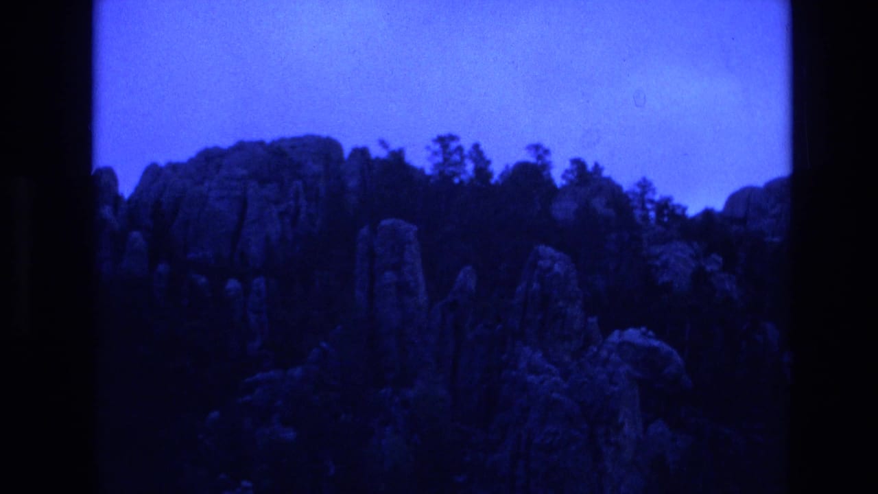 1974 South Dakota 8mm frame showing rocky formations and dark trees along a ridge beneath a strong blue-tinted sky.