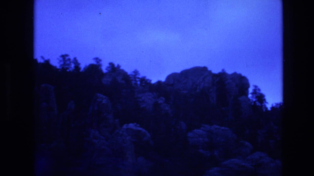 1974 South Dakota home-movie frame with rugged rock outcrops, silhouetted trees, and a dark contact-sheet divider against a blue cast.