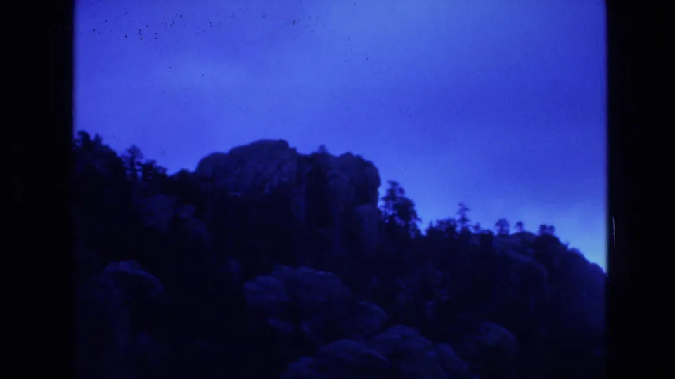 1974 South Dakota 8mm frame showing a darker section of rocky hillside with trees silhouetted against the blue-shifted sky.