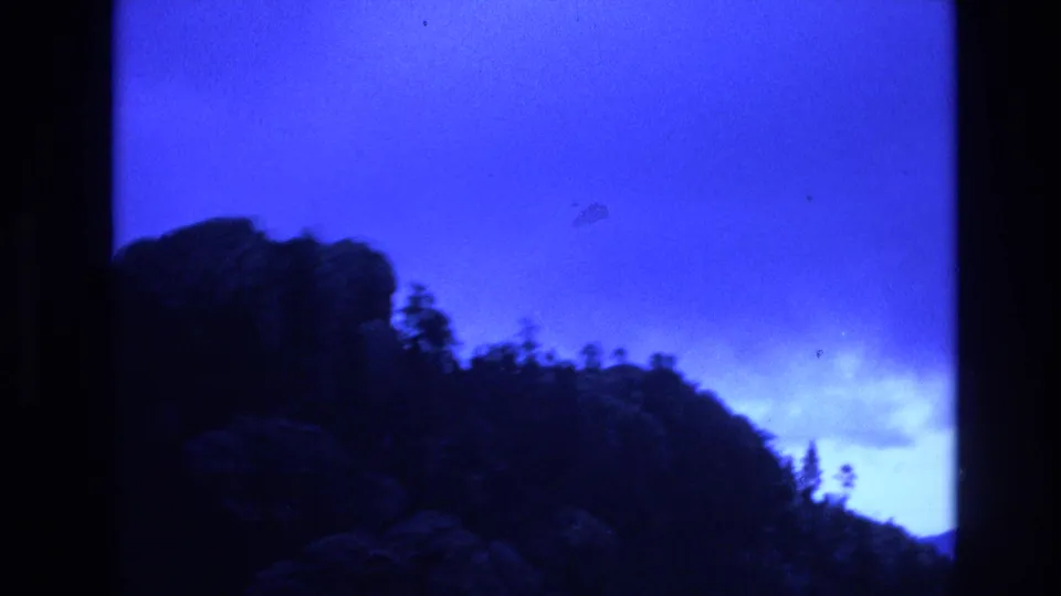 1974 South Dakota home-movie frame showing a rocky, tree-covered slope with blue sky and pale cloud cover near the horizon.