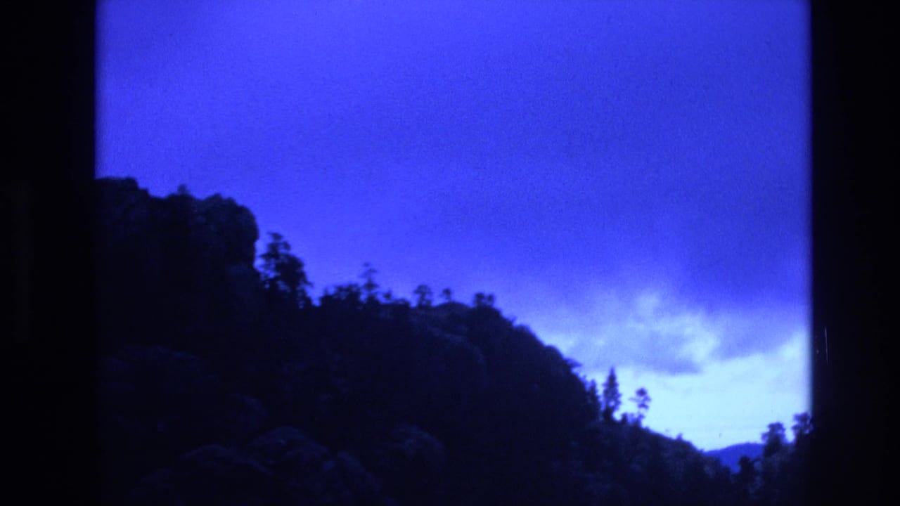 1974 South Dakota 8mm frame showing a dark wooded ridge and rocky hillside silhouetted against an intense blue dusk sky with low clouds.
