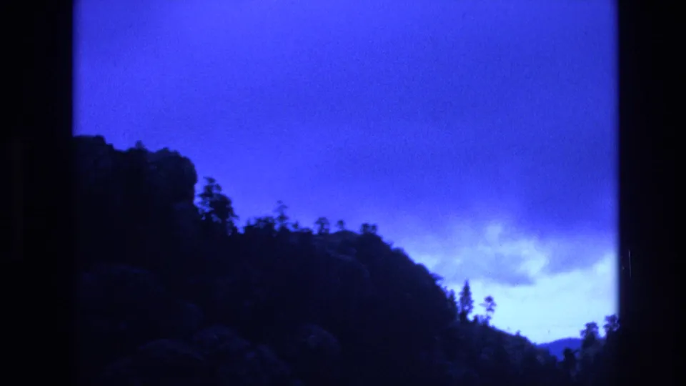 1974 South Dakota 8mm frame showing a dark wooded ridge and rocky hillside silhouetted against an intense blue dusk sky with low clouds.