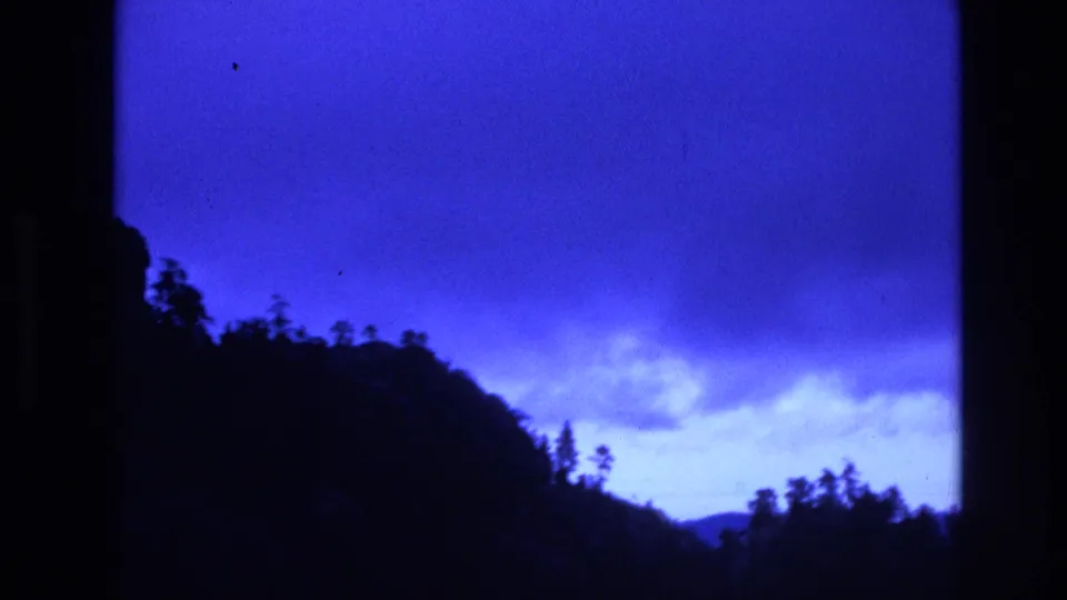 1974 South Dakota 8mm frame with a dark forested ridge rising across the lower frame under a saturated blue sky and pale cloud bank.