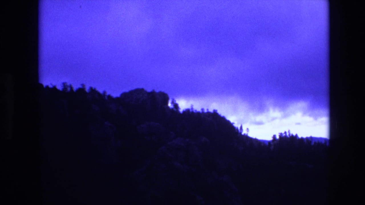 1974 South Dakota 8mm frame showing a silhouetted wooded ridge and rocky hillside under a deep blue, overcast sky.