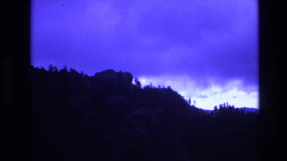 1974 South Dakota 8mm frame showing a silhouetted wooded ridge and rocky hillside under a deep blue, overcast sky.