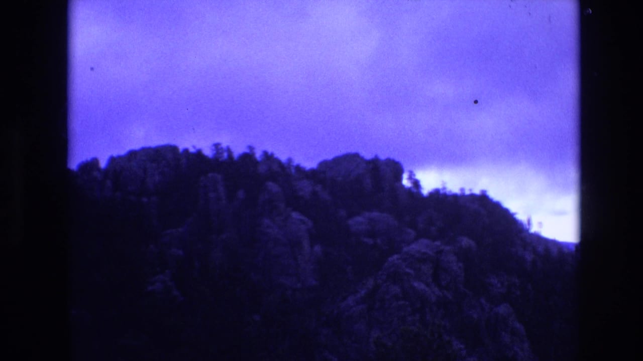 1974 South Dakota 8mm frame with rocky wooded outcrops and a dark ridgeline set against a blue clouded sky.