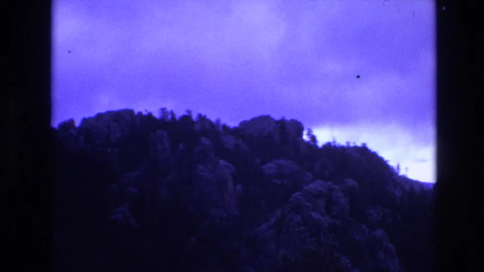 1974 South Dakota 8mm frame with rocky wooded outcrops and a dark ridgeline set against a blue clouded sky.