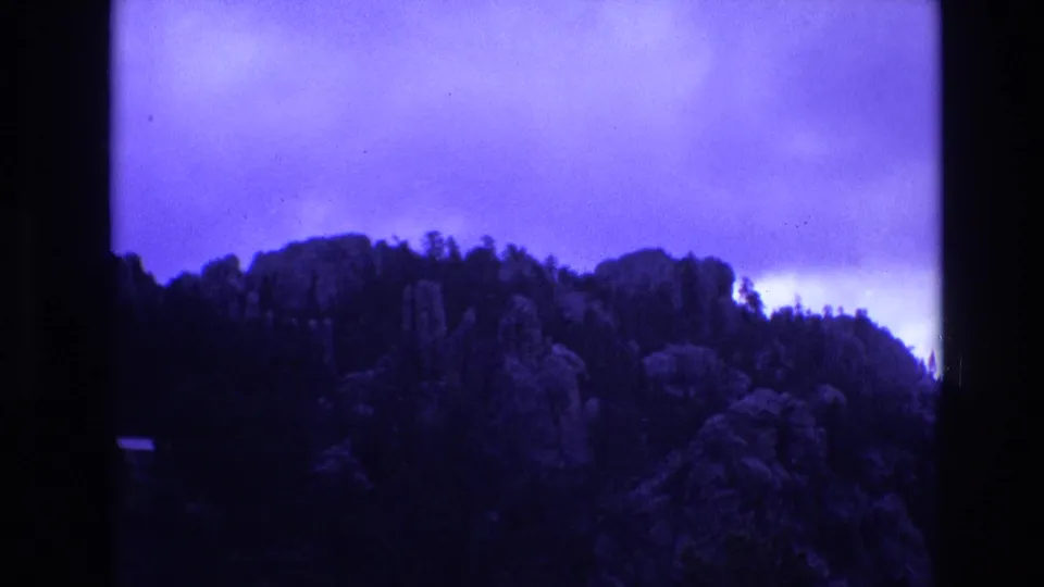 1974 South Dakota 8mm frame showing a wooded mountain ridge with rocky forms beneath a bright band of blue overcast sky.