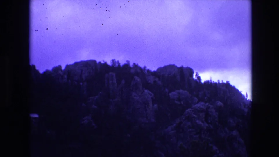 1974 South Dakota 8mm frame with a blue-tinted wooded and rocky ridge, soft grain, and clouded sky filling the upper frame.
