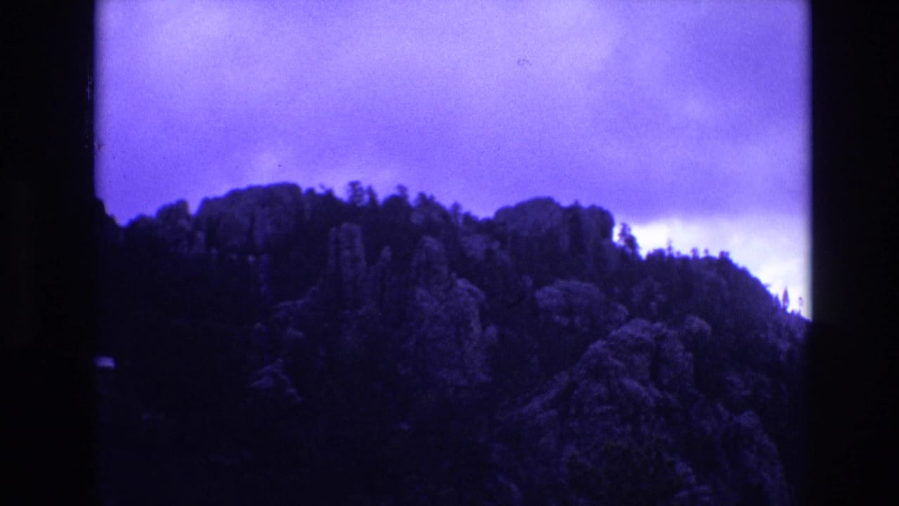 1974 South Dakota 8mm frame showing a forested rocky ridgeline under a bright overcast sky with dark film-edge borders and purple color shift.