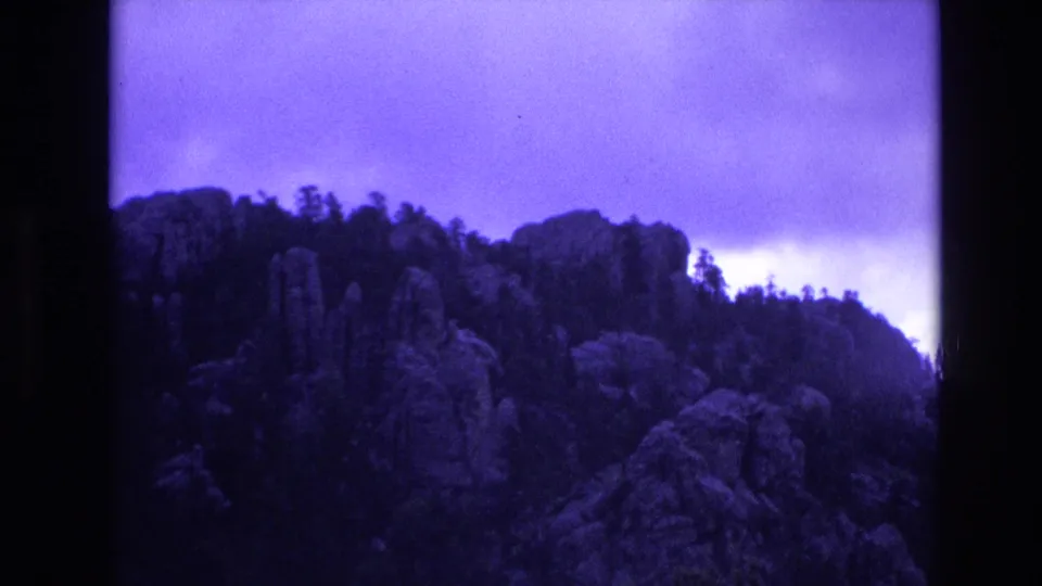 1974 South Dakota 8mm frame with rugged rock outcrops, dark tree silhouettes, and an overcast sky affected by a strong purple-blue film cast.