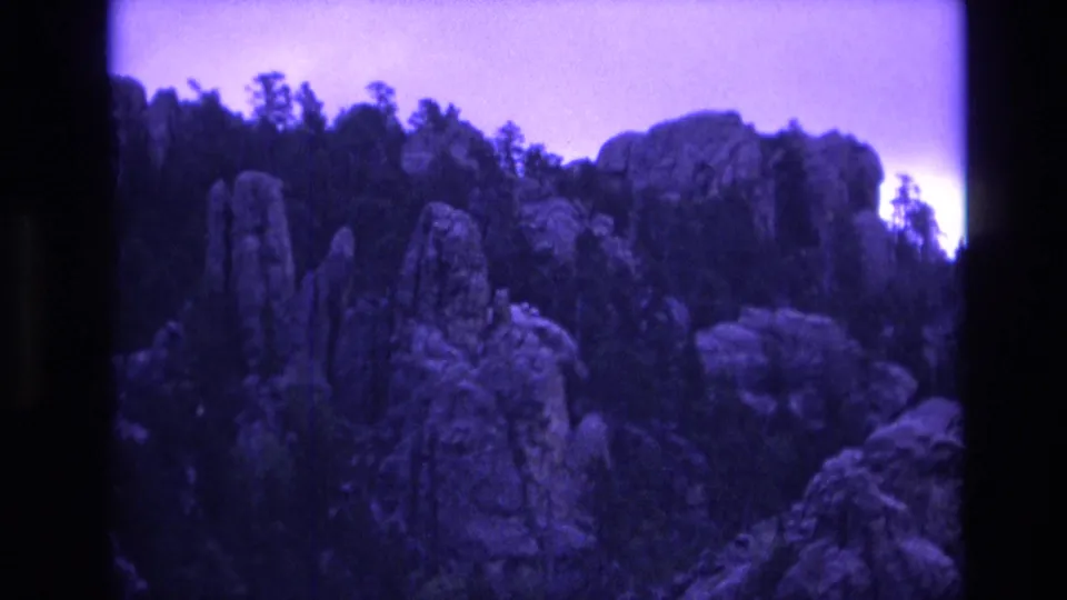 1974 South Dakota 8mm frame showing taller rocky formations and surrounding forest, with soft focus, visible grain, and purple-tinted daylight exposure.