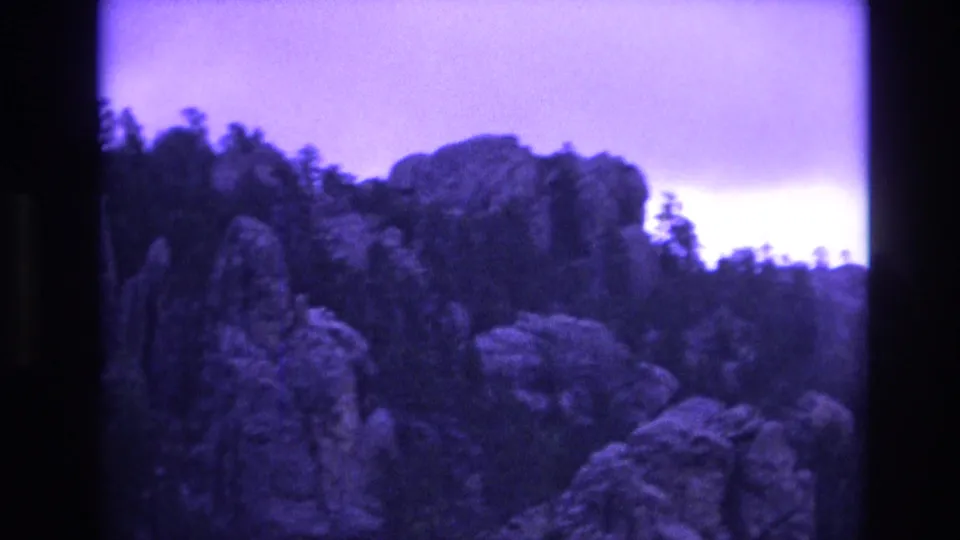 1974 South Dakota 8mm frame of a rocky, tree-covered slope beneath a bright overcast band, framed by dark gate edges and heavy purple color shift.
