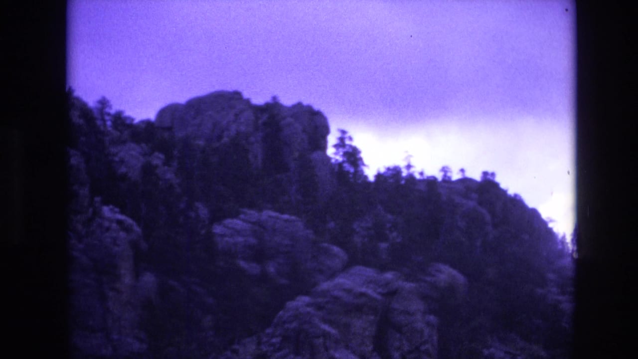 1974 South Dakota 8mm frame showing a rocky ridge with evergreen silhouettes under a purple-tinted storm sky.