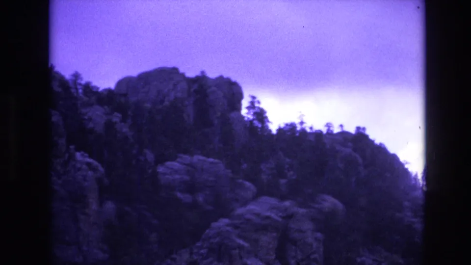1974 South Dakota 8mm frame showing a rocky ridge with evergreen silhouettes under a purple-tinted storm sky.
