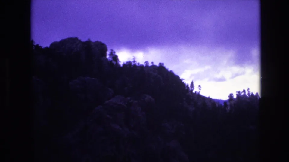 1974 South Dakota 8mm frame showing a rugged, tree-lined hillside beneath a bright opening in heavy purple-tinted clouds.