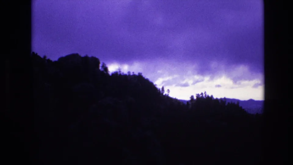 1974 South Dakota home-movie frame with a forested ridge in silhouette, distant hills, and a broad purple storm cloud bank overhead.