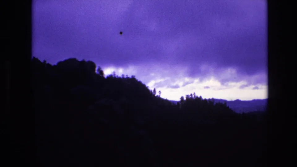 South Dakota 1974 8mm frame showing a dark wooded ridgeline beneath a purple twilight sky with layered clouds and low-light grain.