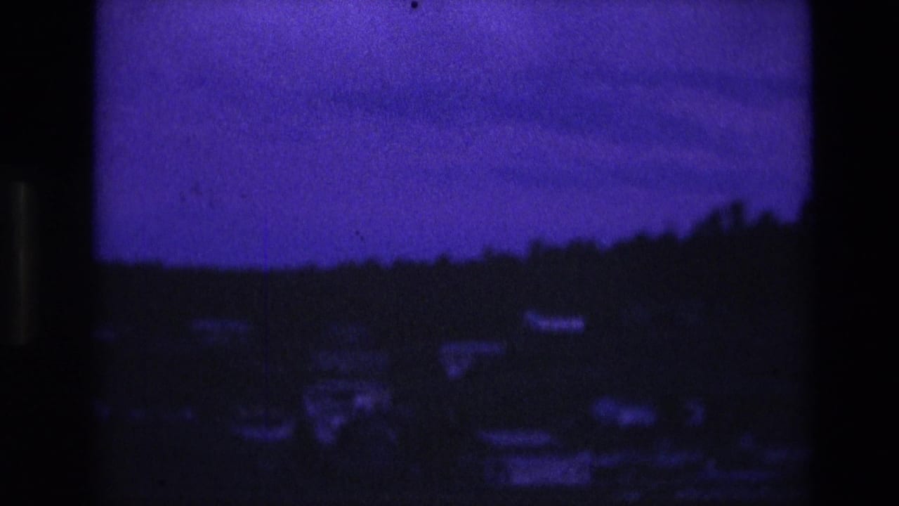 1974 South Dakota 8mm frame showing a silhouetted wooded hill and faint scattered houses beneath a low blue-purple dusk sky.