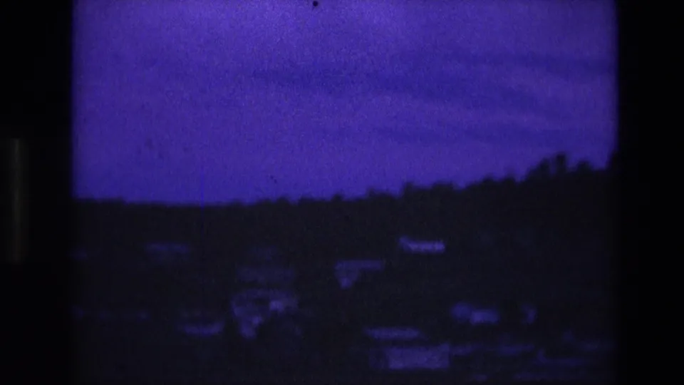1974 South Dakota 8mm frame showing a silhouetted wooded hill and faint scattered houses beneath a low blue-purple dusk sky.