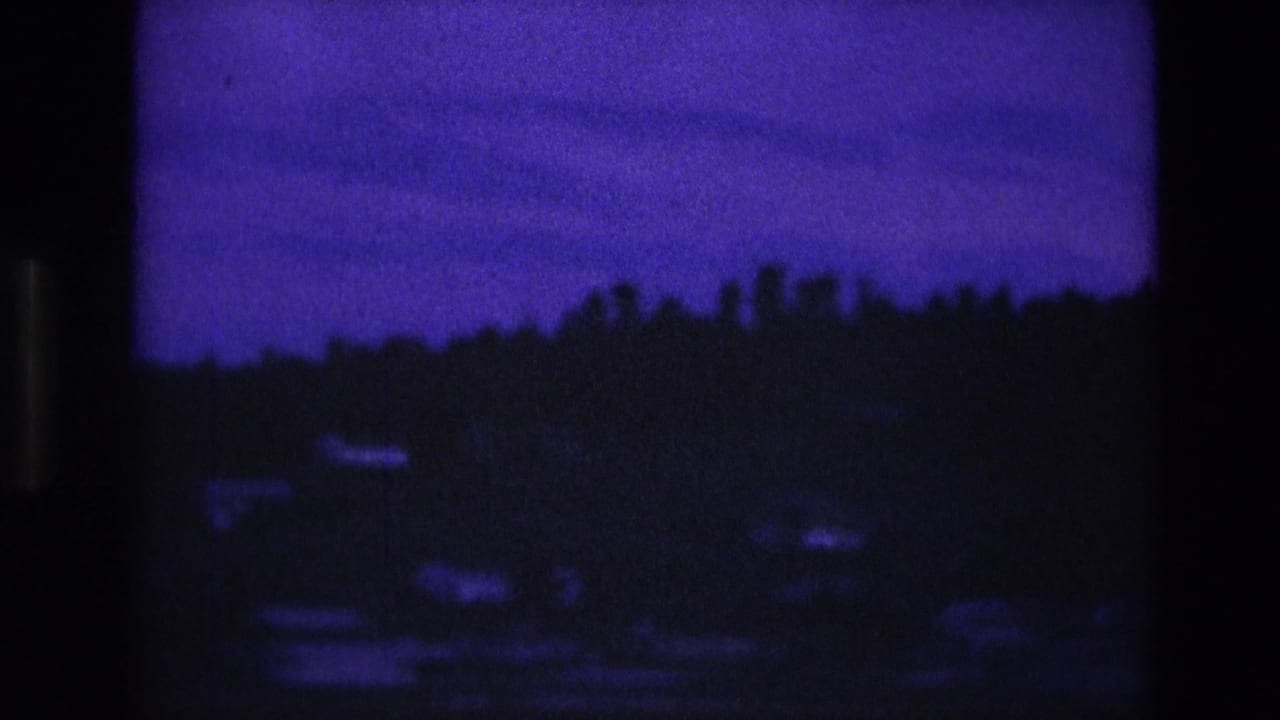 1974 South Dakota home-movie frame with a dark tree-covered ridge and small pale houses softened by low-light grain and blue color shift.