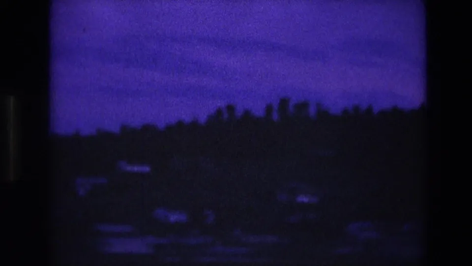 1974 South Dakota home-movie frame with a dark tree-covered ridge and small pale houses softened by low-light grain and blue color shift.