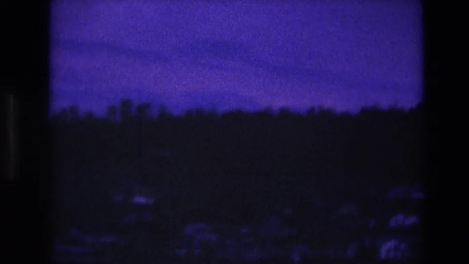 1974 South Dakota 8mm frame showing a shadowed wooded slope with dim residential shapes and a grainy purple-blue evening sky.