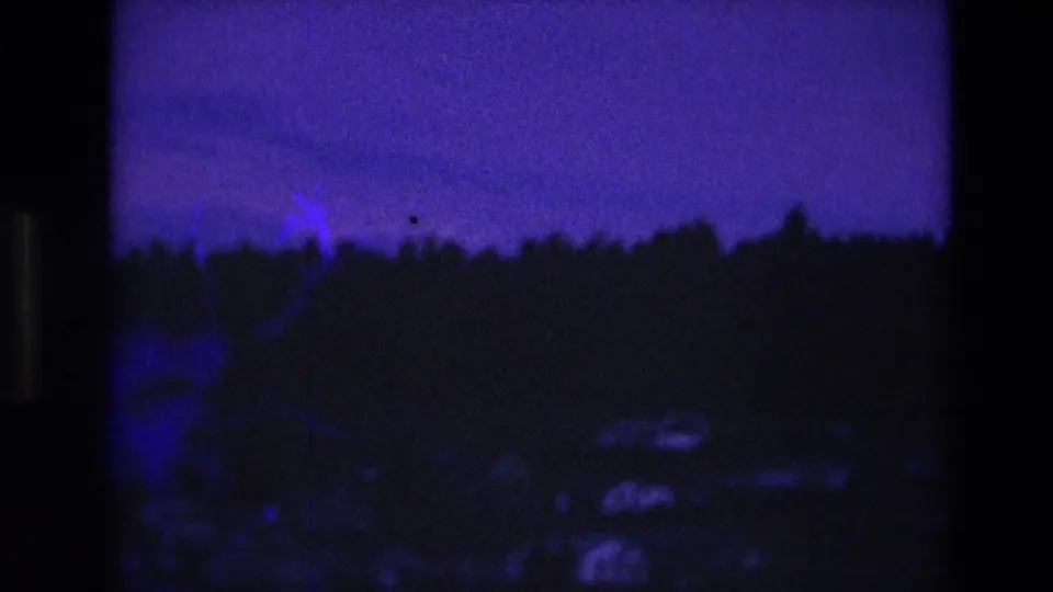 1974 South Dakota home-movie frame with a dark wooded hillside, distant houses, and a blue flare or exposure mark near the tree line.