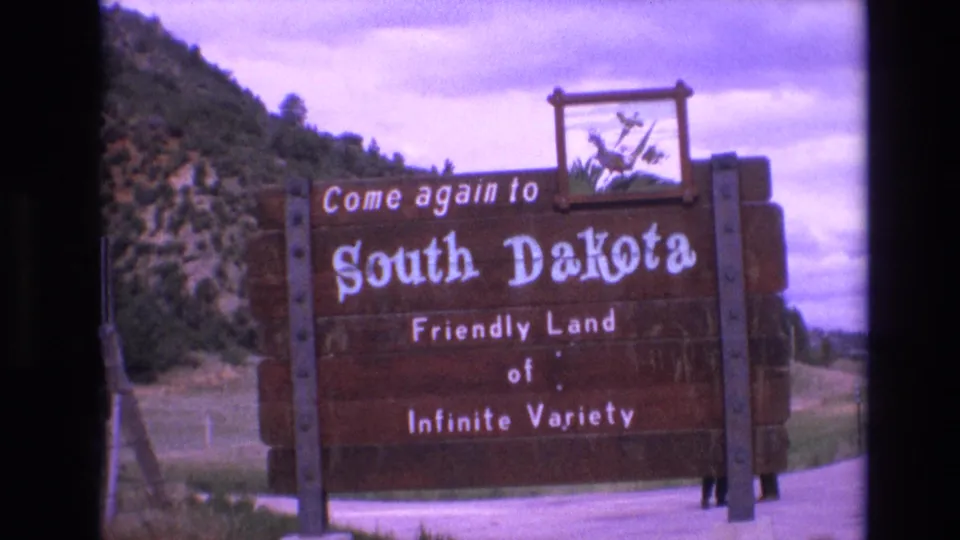 1974 South Dakota 8mm frame showing a wooden roadside sign reading Come again to South Dakota, Friendly Land of Infinite Variety, with a hillside and cloudy sky behind it.