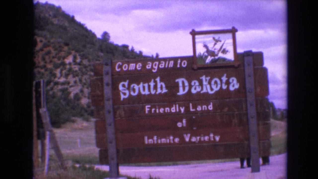 Wooden South Dakota welcome sign beside a road with hills and cloudy sky behind it.
