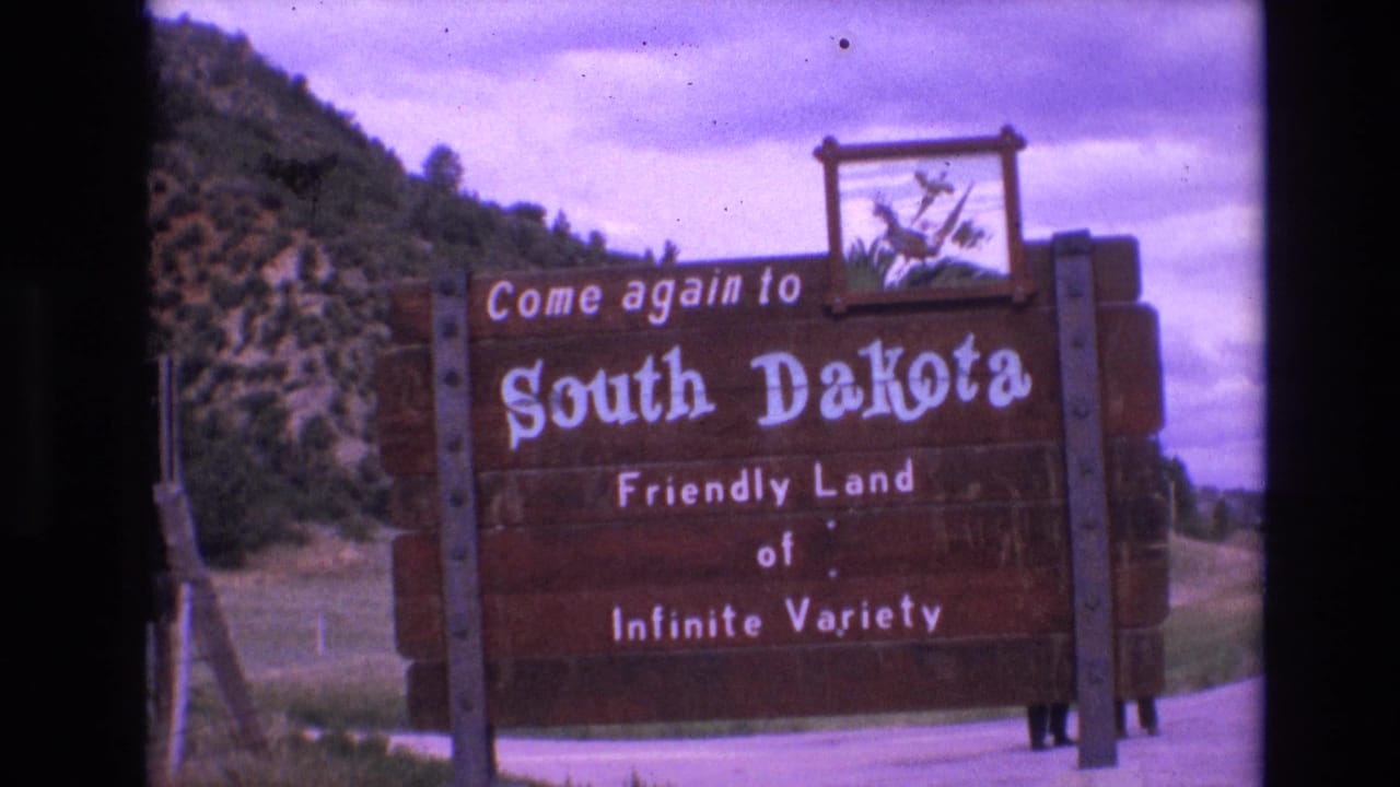 South Dakota wooden sign fills the frame with hillside, road, and cloudy sky visible around it.