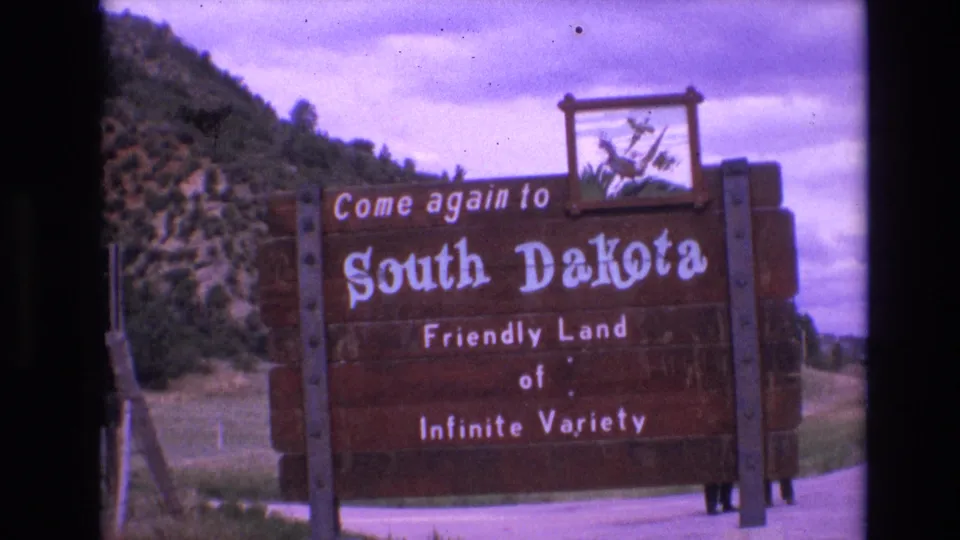 1974 South Dakota travel footage frame showing the wooden welcome sign and prairie chicken emblem beside a rural road under a purple-tinged cloudy sky.