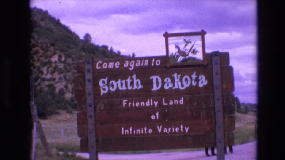 1974 8mm frame holding on the South Dakota welcome sign, with the slogan “Friendly Land of Infinite Variety” visible against a rural hillside backdrop.