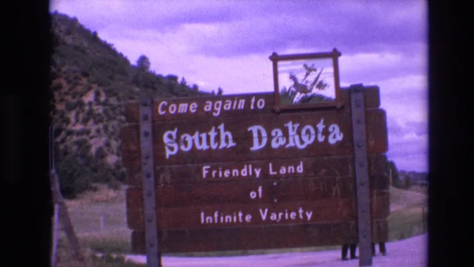 Final 1974 8mm frame of the South Dakota roadside welcome sign, with its painted text and emblem framed against hills and an overcast purple-toned sky.