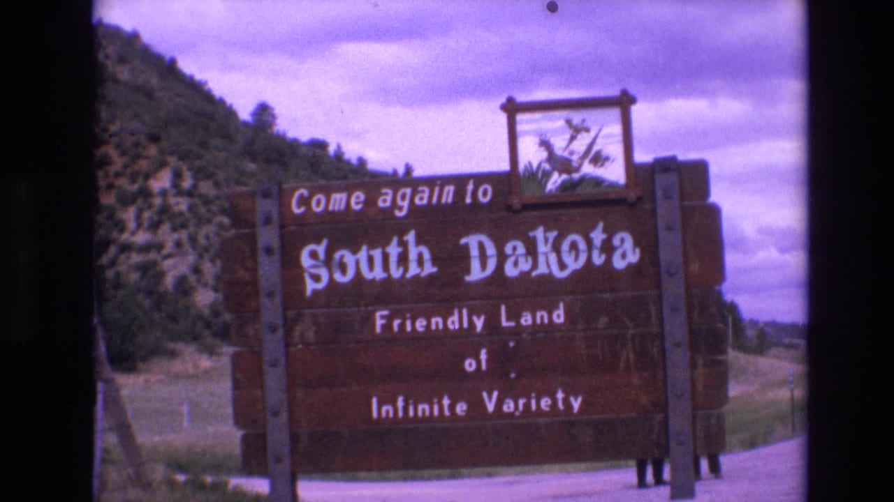 1974 8mm frame of a wooden South Dakota welcome sign reading “Come again to South Dakota” beside a rural roadside with hills, trees, and cloudy sky.