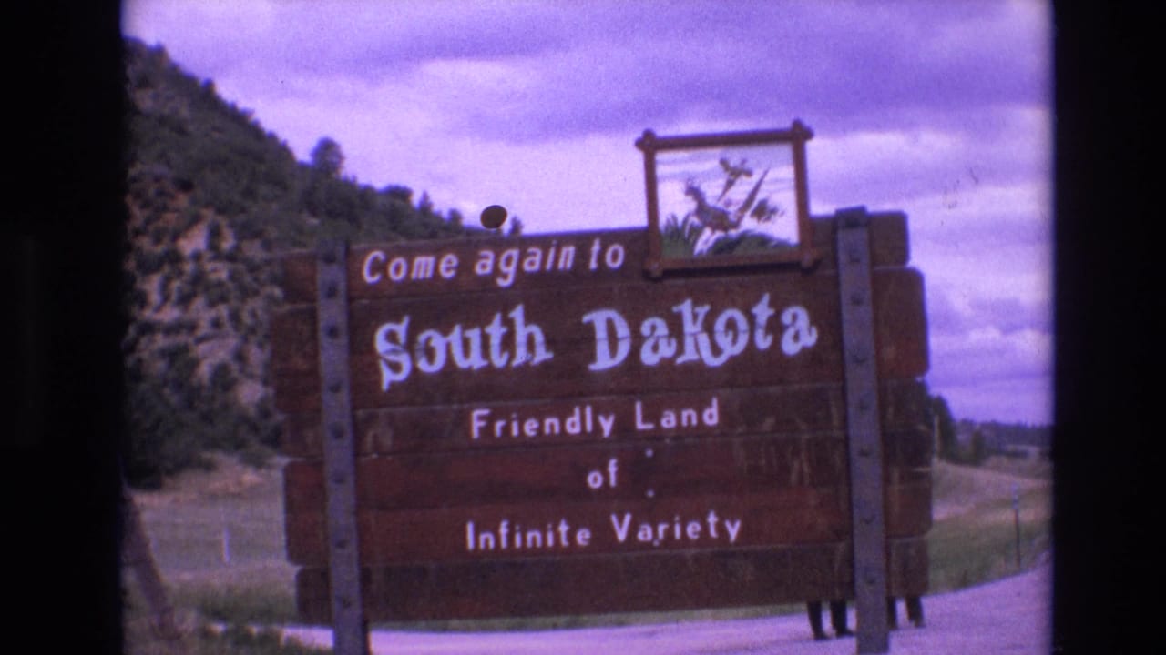 South Dakota welcome sign framed closer, with cloudy sky and hillside behind it.