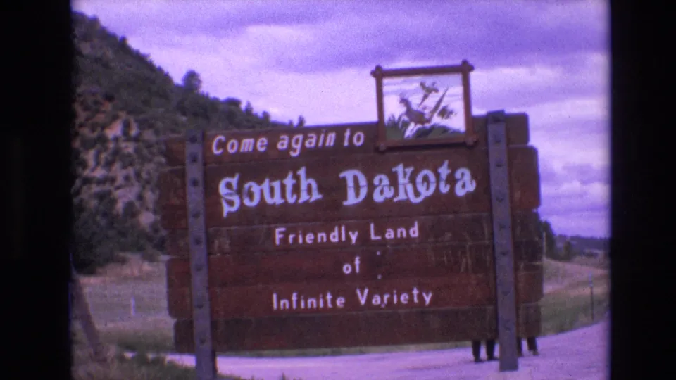 1974 home-movie frame of the South Dakota border welcome sign, with the slogan visible against wooden boards and a softly focused rural landscape behind it.