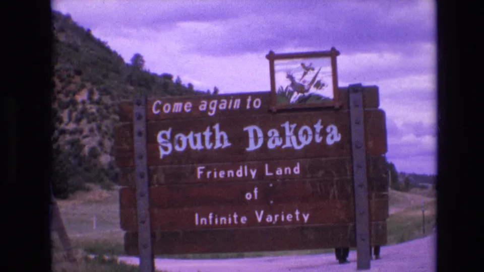 Final 1974 8mm frame of the wooden South Dakota welcome sign beside the roadway, showing the emblem panel, white lettering, hillside, trees, and overcast sky.