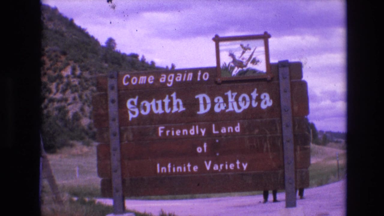 1974 8mm frame showing a large wooden South Dakota welcome sign reading Come again to South Dakota, with hillside, trees, cloudy sky, and partial people behind the sign.