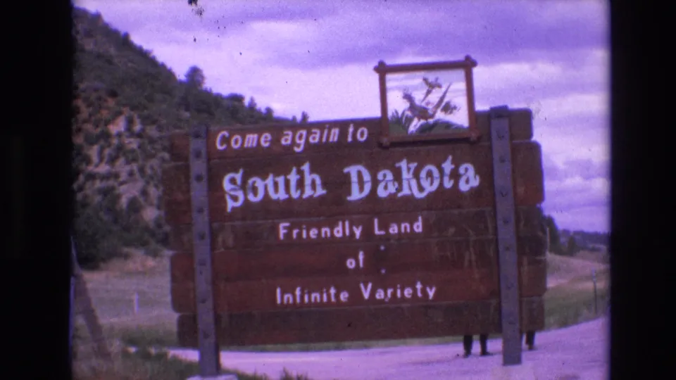 1974 8mm frame showing a large wooden South Dakota welcome sign reading Come again to South Dakota, with hillside, trees, cloudy sky, and partial people behind the sign.