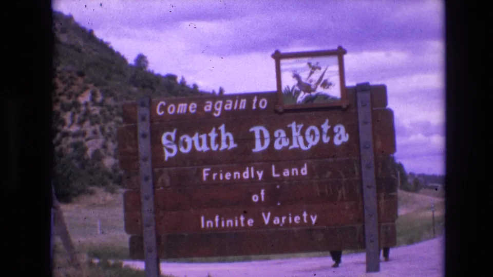 1974 South Dakota travel footage frame with the wooden welcome sign centered near a rural road, backed by a green hillside, trees, and overcast sky.