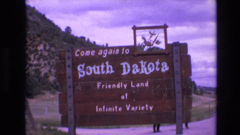 1974 8mm frame of the South Dakota welcome sign reading Friendly Land of Infinite Variety, with grass, road edge, hillside vegetation, and partial people visible below the sign.