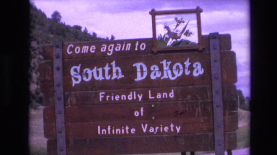 End frame from 1974 8mm footage showing a closer view of the wooden South Dakota welcome sign, its bird emblem, painted lettering, roadside setting, and cloudy sky.