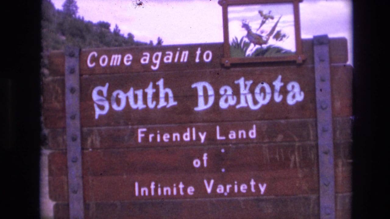 Wooden sign reading Come again to South Dakota with roadside greenery behind it.