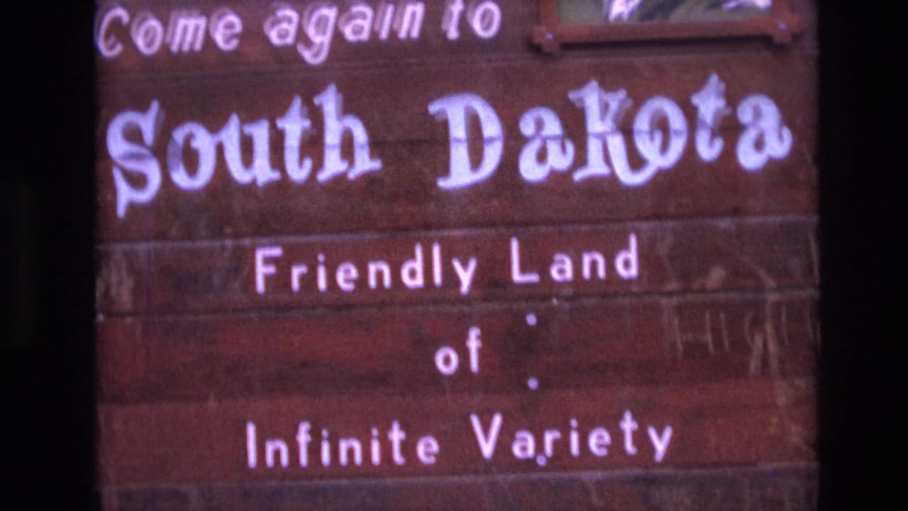 Close 1974 8mm view of the South Dakota welcome sign, emphasizing the painted white lettering and the phrase Friendly Land of Infinite Variety.